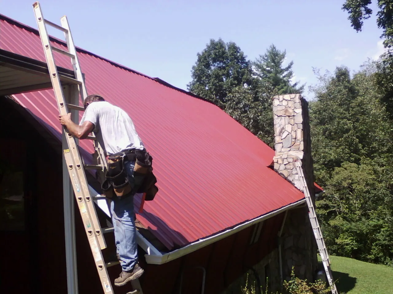 Roofer on a ladder beside a red metal roof and stone chimney — Soffit & Fascia Repair by Roofing Brothers in Port Ewen